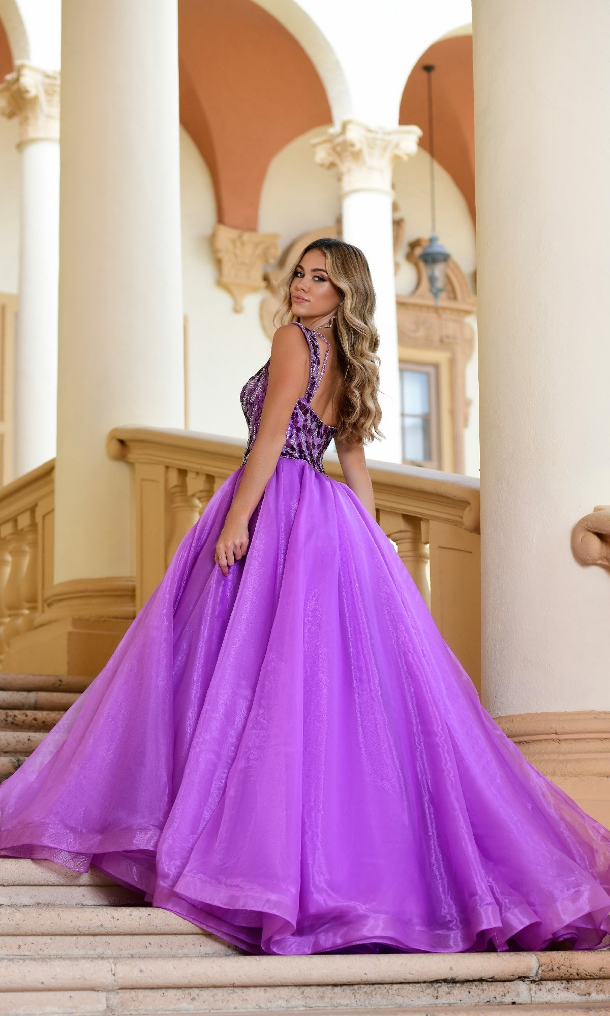 A woman wearing the Long Formal Dress 28579 by Ava Presley, featuring an embroidered bodice and v-neckline, stands on a staircase in an elegant architectural setting, looking back over her shoulder.